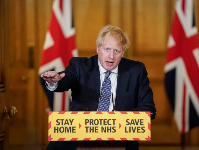 Boris Johnson behind a podium reading 'Stay home, protect the NHS, save lives'