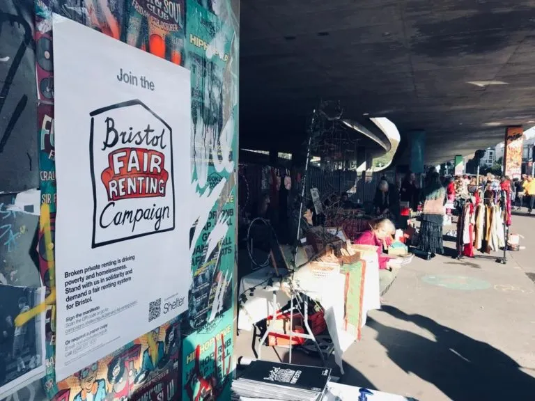 A close up of a poster reading 'Bristol Fair Renting campaign' on an outside wall, with street stalls in the background