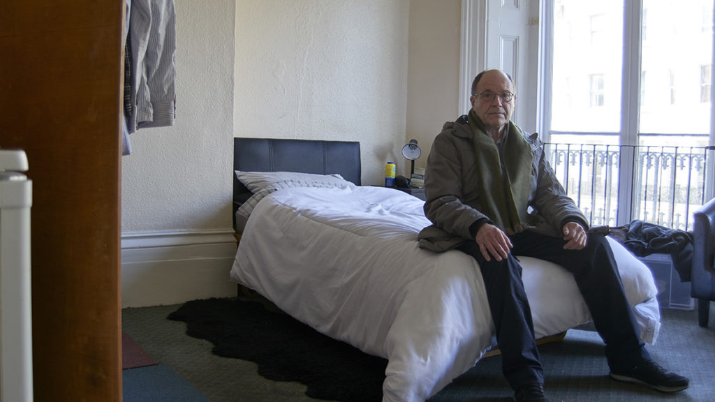 A photograph of an older man sitting on a bed in temporary housing.