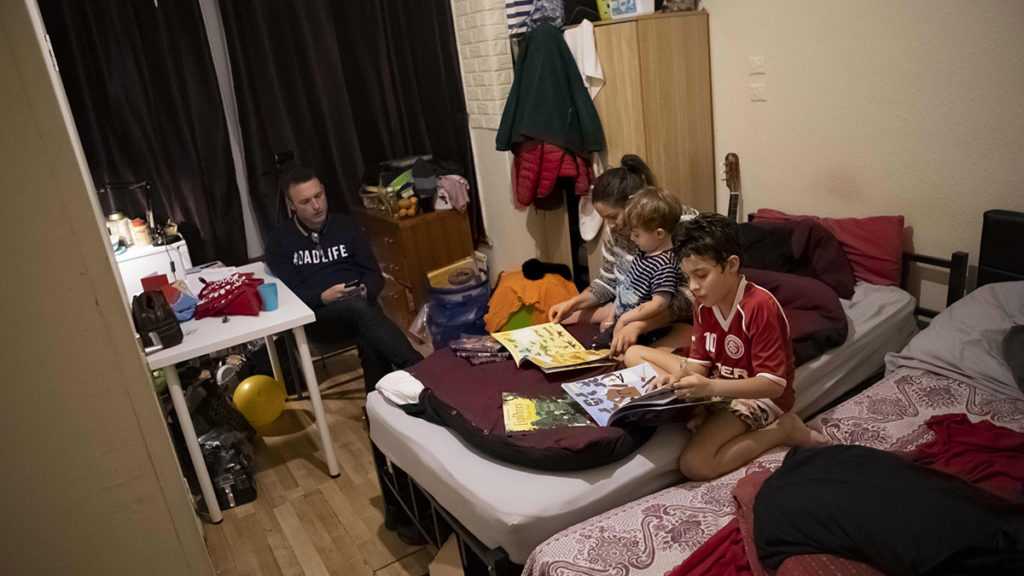 A group of children in a temporary accommodation sit reading on a bed together while their father watches.