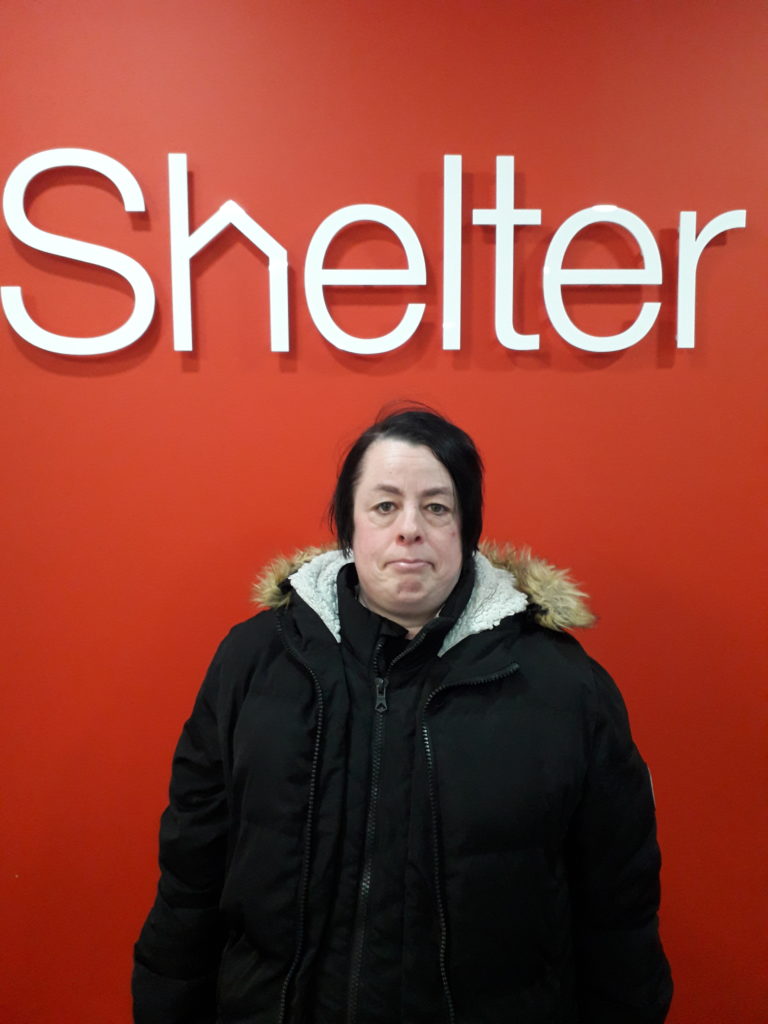 Shelter peer mentor Barbara stands below a red Shelter branded wall.