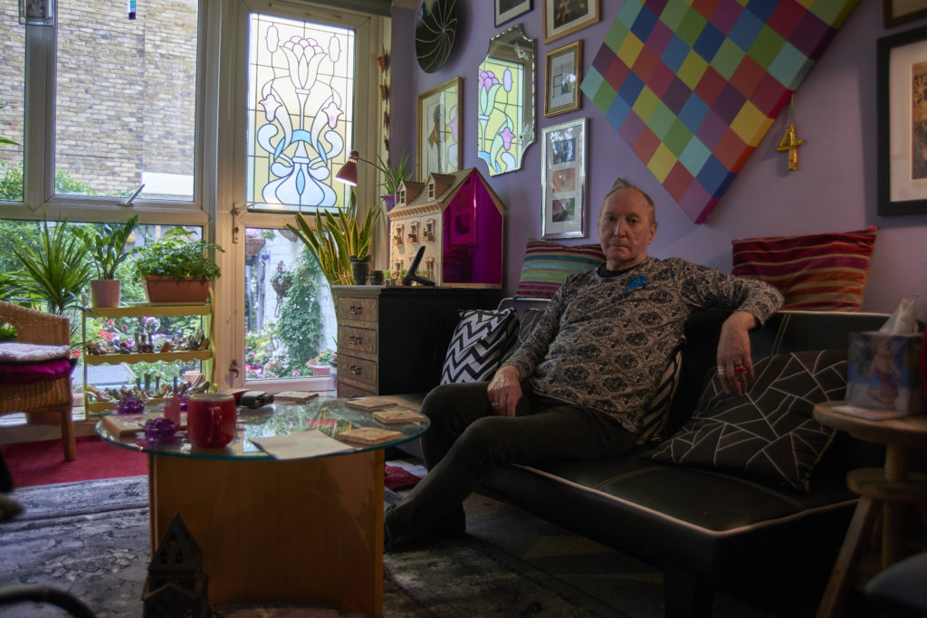 A man in a patterned jumper sitting on a leather sofa in a room of colourful pictures and plants