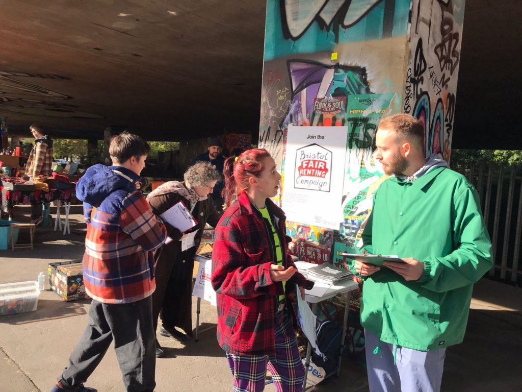 A man in a green jacket and a woman with red hair talking next to a stall set up outside with leaflets and posters