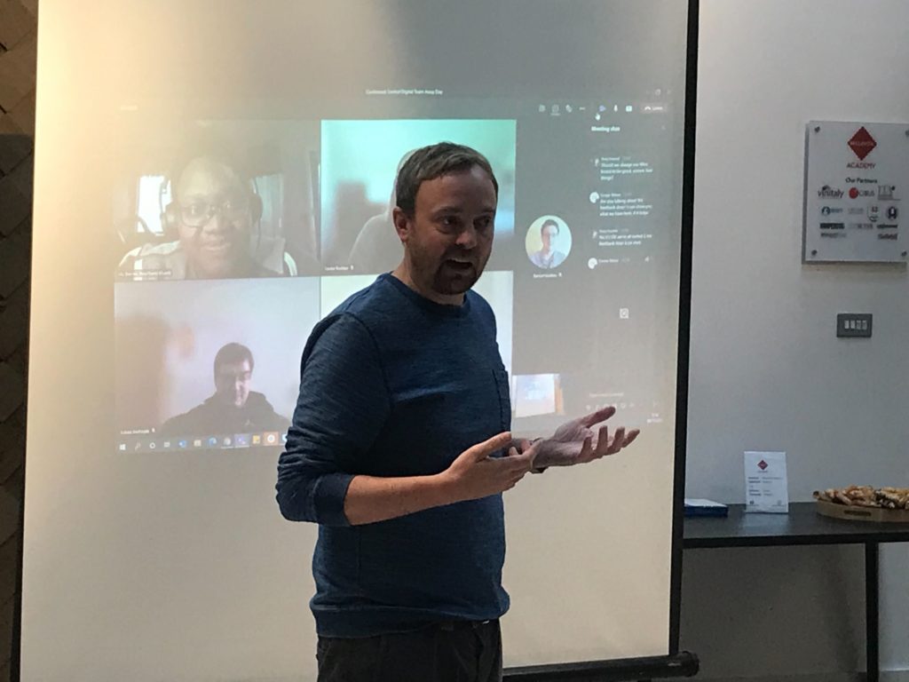 A man presenting in front of a big screen showing a video call