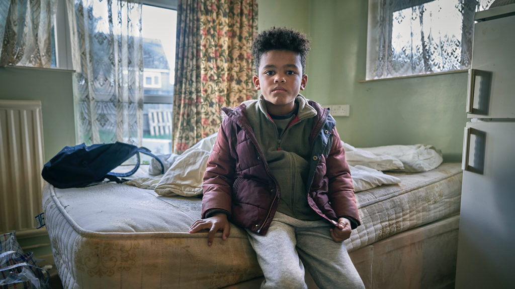 A child in temporary accommodation, sitting on a dirty mattress next to a fridge