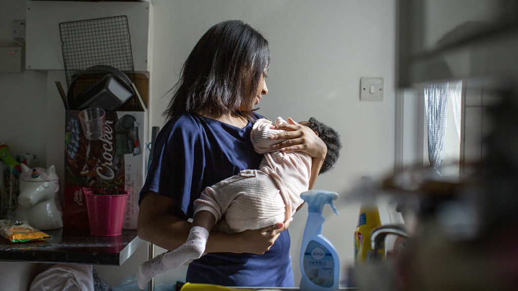 A mother stands in a kitchen holding her sleeping baby