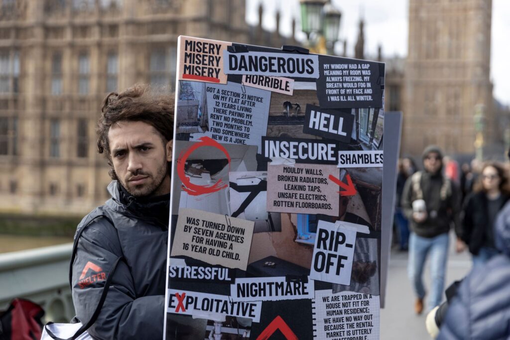 A man in a black, Shelter-branded jacket standing outside the House of Parliament in London, holding a placard with a collage of words and images relating to renting on it.