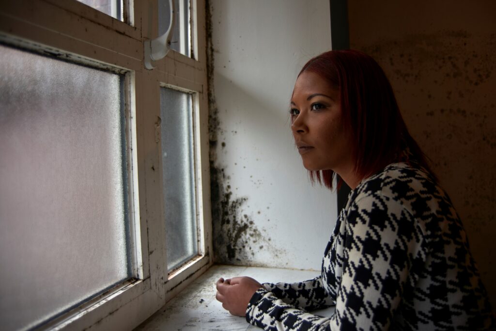 An image of a woman wearing a black and white checked sweater looking out of a dirty window, with mould growing on the walls beside her