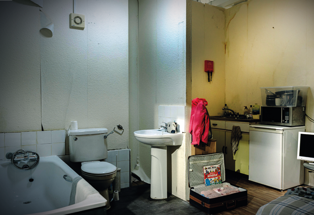 A dirty, damp and cramped bathroom with a packed suitcase open on the floor