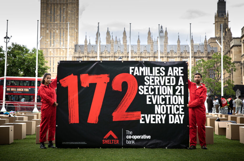 Two campaigners raise a banner outside parliament. It reads: 172 families are served a section 21 eviction notice every day.