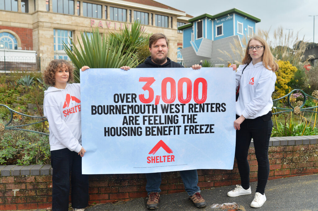 Three people in Shelter tops stand outside a house in Bournemouth. They hold a sign reading 'over 3,000 Bournemouth West renters are feeling the housing benefit freeze'