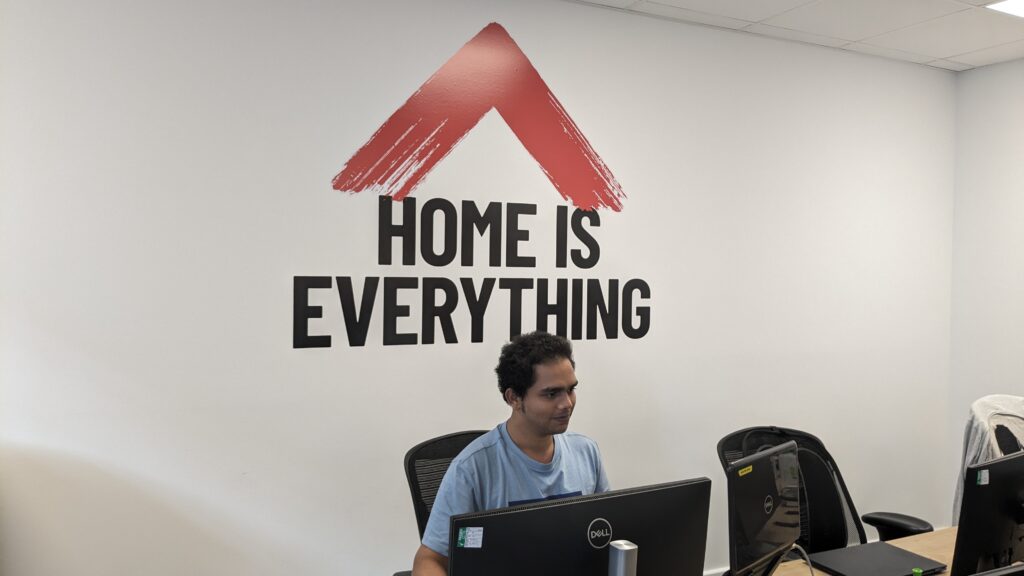 Saurabh sits at a desk with a double screen monitor. The sign on the wall behind him reads 'Home is everything'.